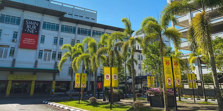 The Swinburne University campus featuring modern buildings partially shaded by tall palm trees in the foreground. Clear skies and open walkways create a welcoming, sunny atmosphere.