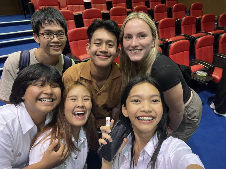 An international student smiling with a group of local Thai students inside a university auditorium, capturing the friendships built through cultural immersion abroad.