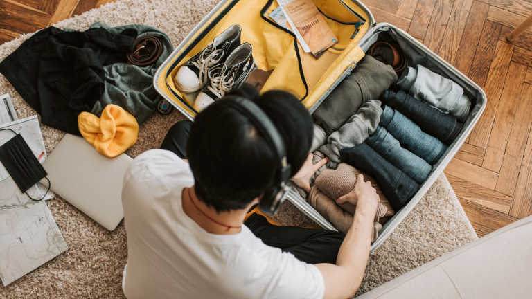 A student wearing headphones packing a neatly organised suitcase on the floor, surrounded by clothes, shoes, a laptop and travel maps, preparing their study abroad packing list.